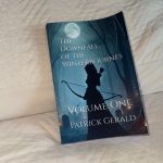 The paperback of The Downfall of the Winterbournes: Volume One by Patrick Gerald, displayed on a bed. The book cover features a silhouetted figure with a bow in a dark, snowy landscape, representing the dark themes of betrayal, survival, and family drama in the medieval fantasy series.